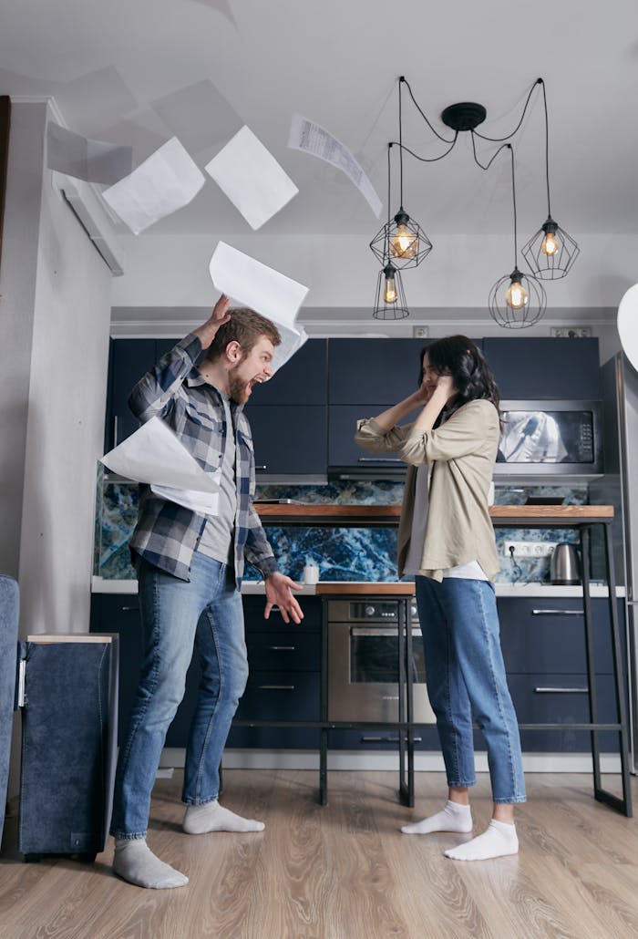 A man and woman argue in a kitchen with papers flying around.